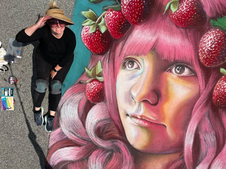 Jessi in a sun hat beside her chalk portrait of a pink-haired girl crowned with strawberries