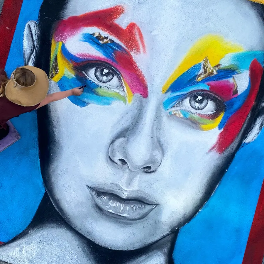 Jessi in a sun hat painting bold red, yellow and blue strokes across a chalk portrait’s eyes