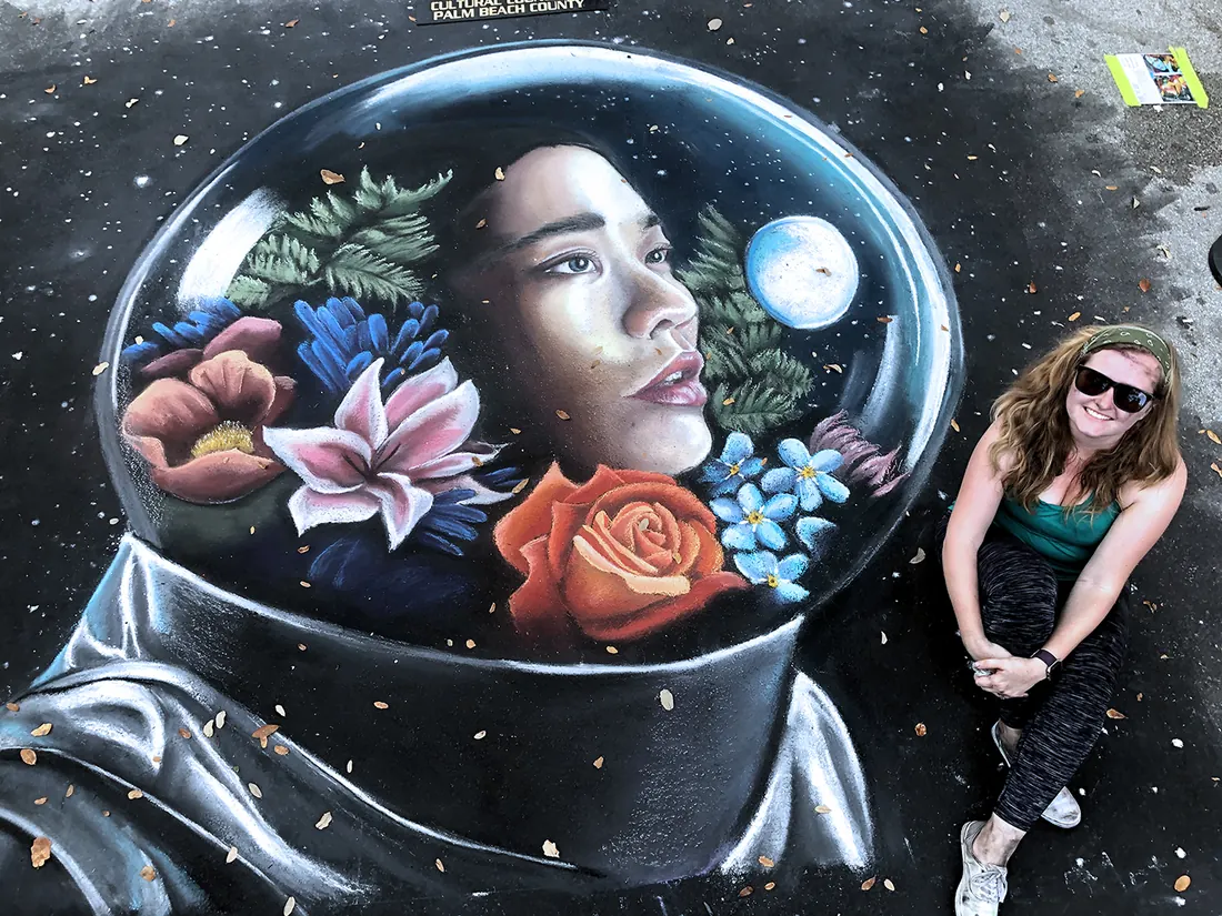 Jessi kneeling beside her street chalk portrait of a woman inside an astronaut helmet filled with flowers, Lake Worth 2019