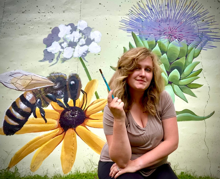 Jessi holding a paintbrush in front of her garden wall mural of a bee on a black-eyed Susan