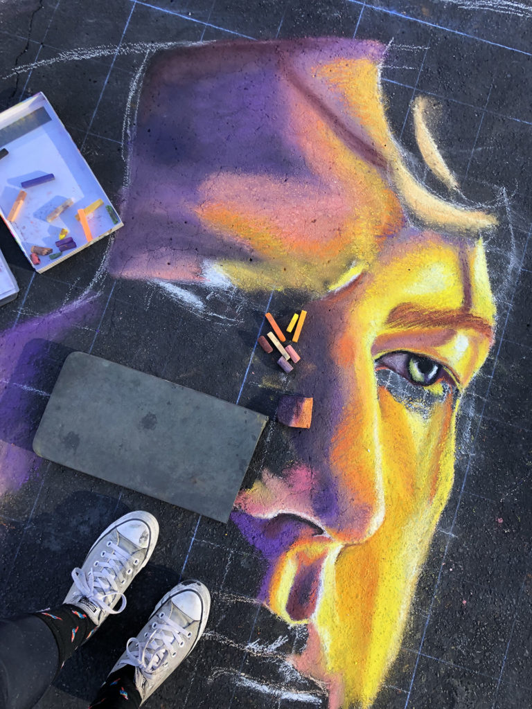 Jessi's sneakers beside an in-progress chalk portrait of a man in purple and gold, from above