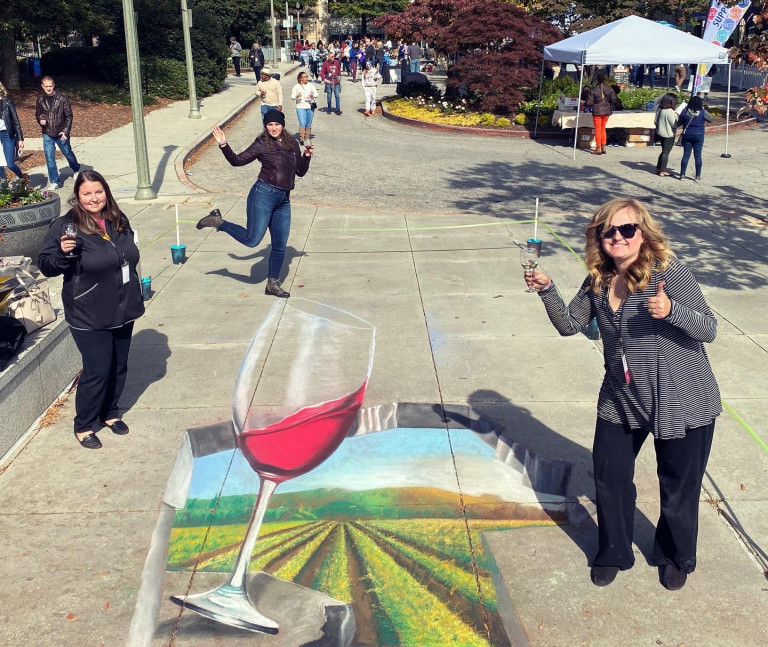 Three people toasting around a 3D chalk wine glass above a vineyard landscape
