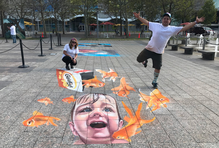 Two people posing on the 3D Goldfish chalk mural of a laughing child and goldfish crackers