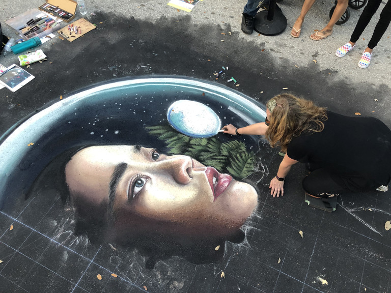 Jessi drawing a chalk portrait of a woman gazing up from inside an astronaut helmet filled with flowers