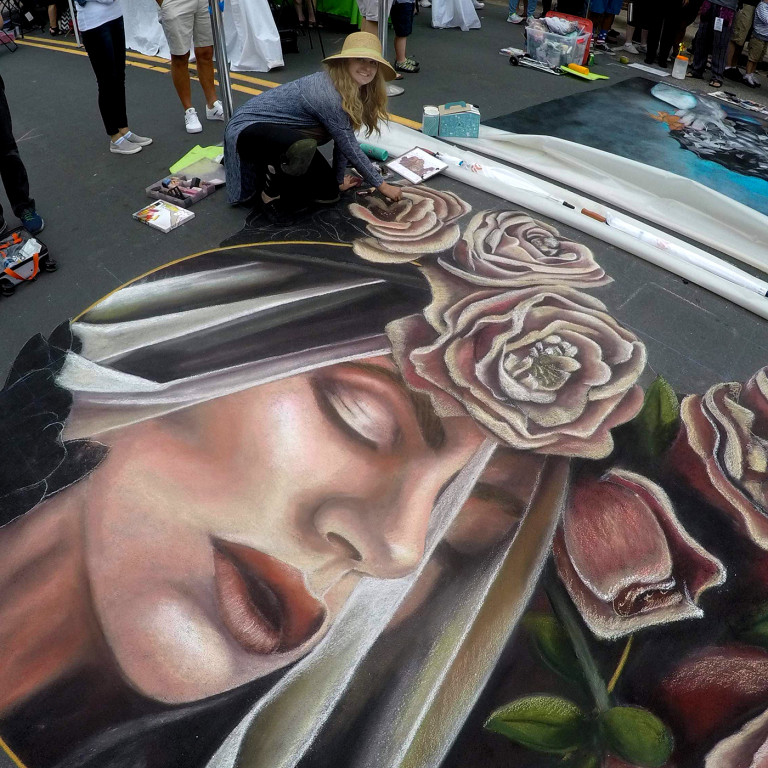 Jessi in a sun hat drawing a chalk portrait of a veiled woman crowned with roses