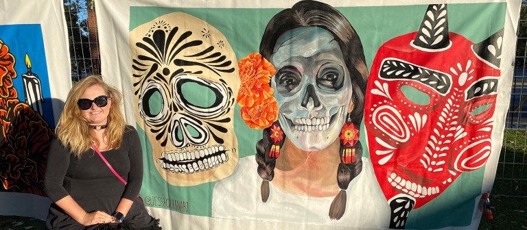 Jessi seated in front of her Dia de los Muertos mural of a skull-faced woman between two masks
