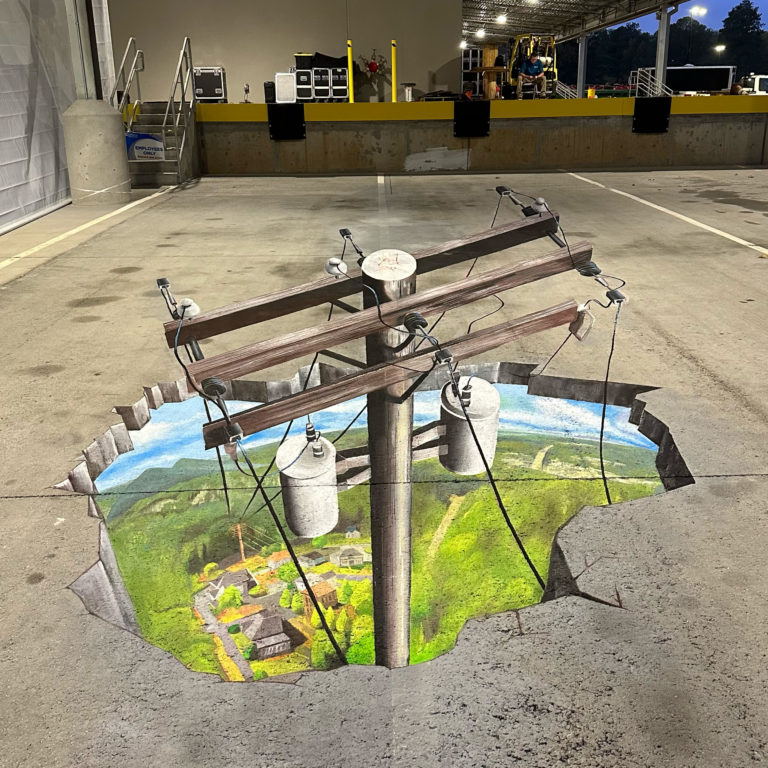 3D chalk mural of a power pole rising out of a hole that reveals green hills and homes below