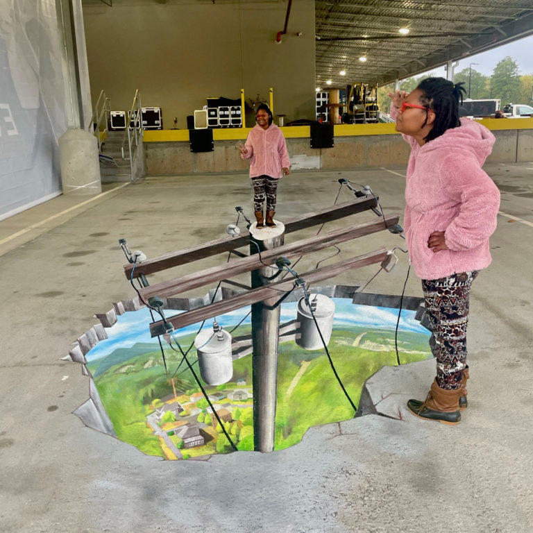 Woman at the edge of the 3D power pole chalk mural while another poses on top of the pole