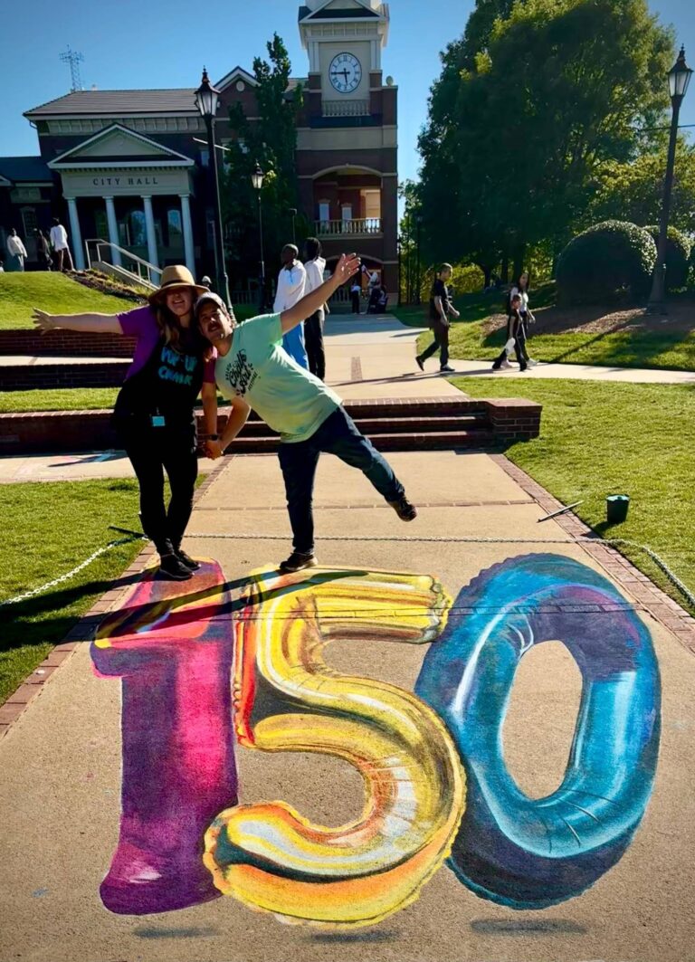 Jessi and a friend posing above giant 3D chalk number balloons reading 150 in front of City Hall