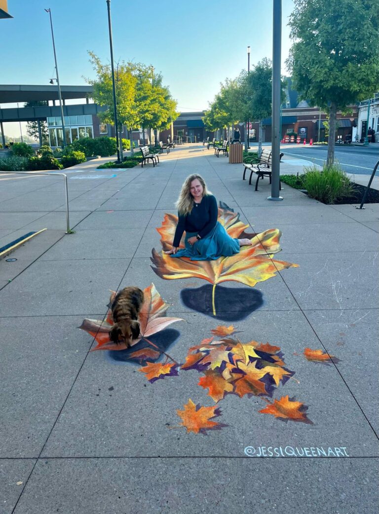 Jessi seated on a giant 3D chalk autumn leaf while her dachshund sniffs a smaller leaf