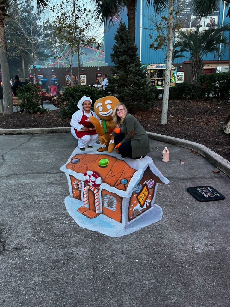 Jessi, a baker, and a gingerbread mascot crouching on top of a 3D chalk gingerbread house