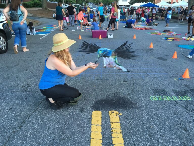Jessi crouching to feed a 3D chalk hummingbird that hovers above the street at a chalk festival