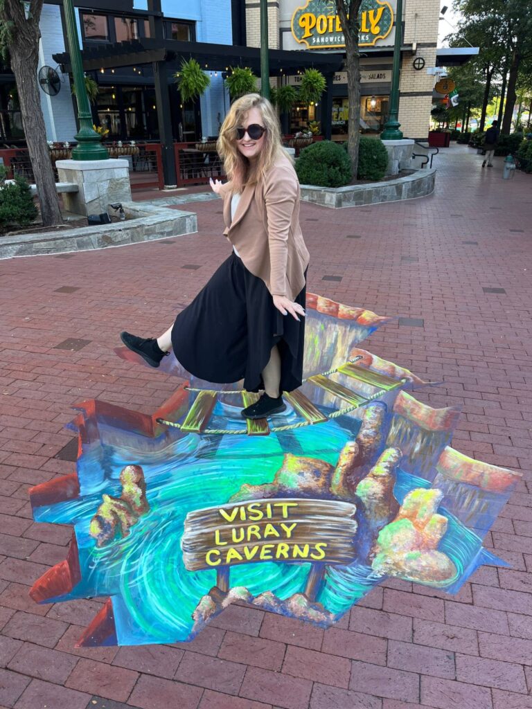 Jessi stepping across a 3D chalk rope bridge over a cavern pool with a Visit Luray Caverns sign