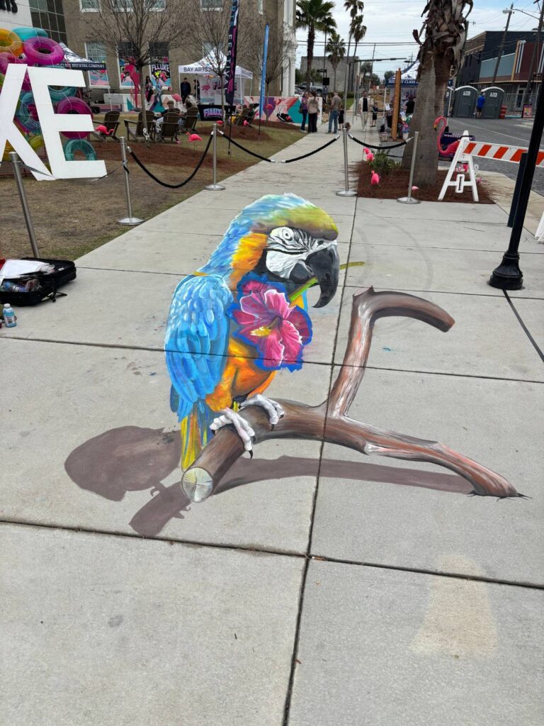 3D chalk blue-and-gold macaw holding a pink hibiscus, perched on a branch at an arts festival