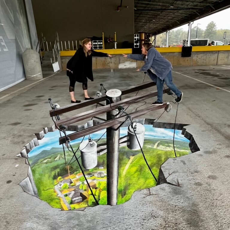 Two women reaching for each other across the crossarms of a 3D chalk power pole above a valley town