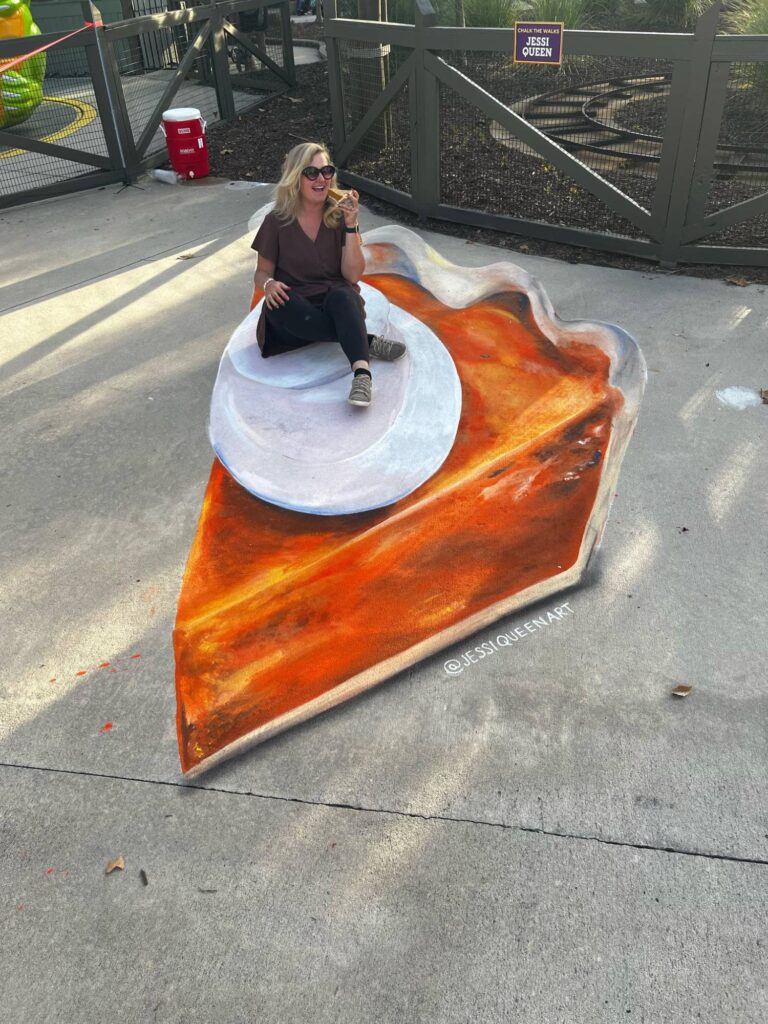 Jessi taking a bite while seated on a giant 3D chalk slice of pumpkin pie with whipped cream
