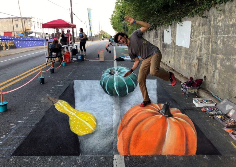 Man balancing on a 3D chalk orange pumpkin beside a green pumpkin and a yellow gourd in a pit, on a closed street during a chalk festival