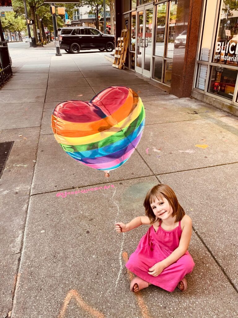 Smiling girl holding the string of a 3D chalk rainbow heart balloon outside Blick Art Materials