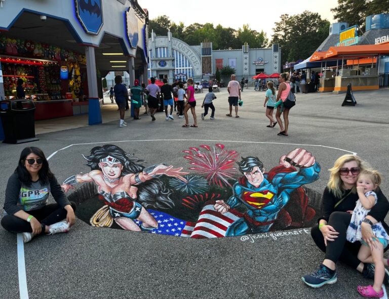 Two women and a toddler seated beside a 3D chalk hole with Wonder Woman and Superman bursting out over fireworks and a flag