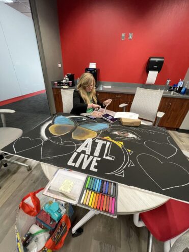 Jessi seated at a table at the ATL Live studio, chalking the blue and yellow butterfly on the ATL Live drawing, with a tray of pastels beside her