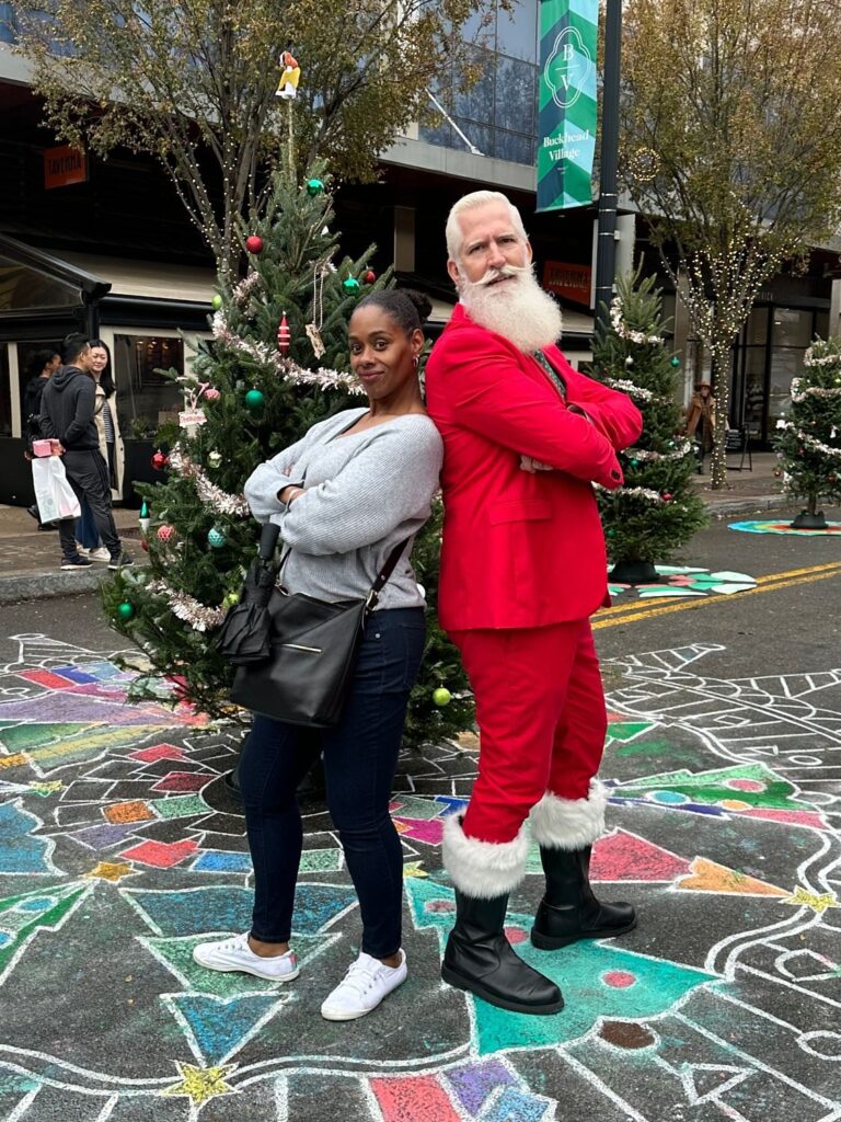A woman and Santa posing back to back on a colorful community chalk mandala in front of a decorated Christmas tree