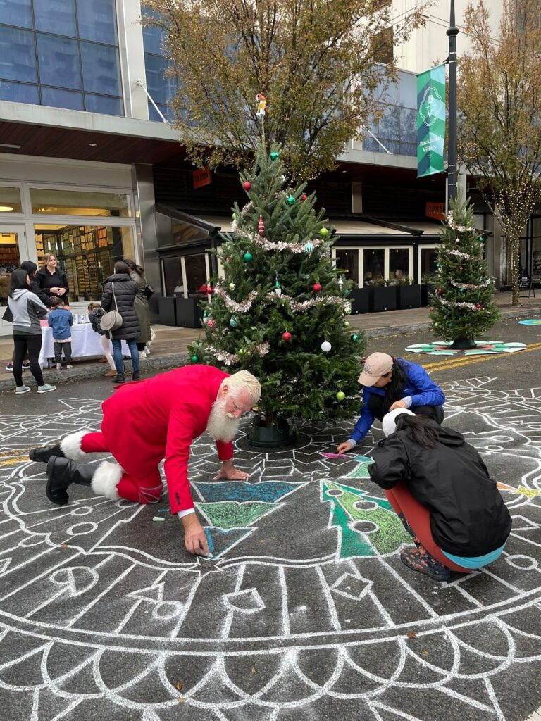 Santa kneeling with kids to color a chalk Christmas tree mandala around a decorated tree at Buckhead Village