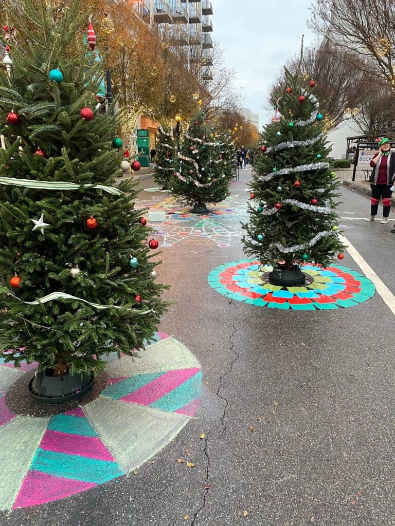 Row of decorated Christmas trees down a closed street, each on a chalk or painted tree skirt in bright patterns