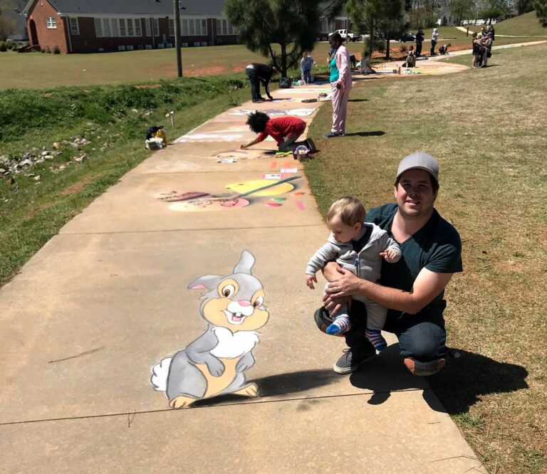 A father and baby crouched beside a chalk drawing of Thumper the bunny on a sidewalk lined with chalk artists