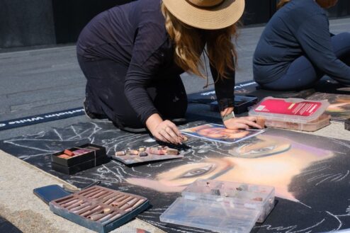 Jessi in a sun hat kneeling over a chalk portrait in progress, holding the reference photo, with trays of pastels beside her