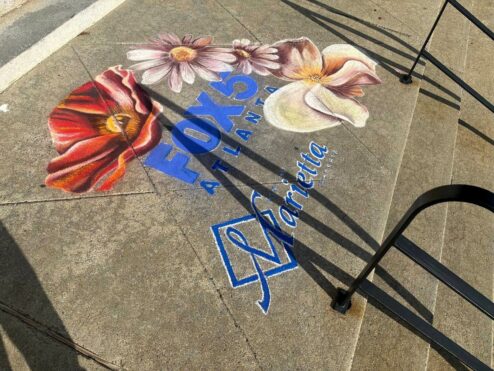 Finished chalk drawing of a red poppy, pink daisies and a white magnolia around the FOX 5 Atlanta and City of Marietta logos in blue