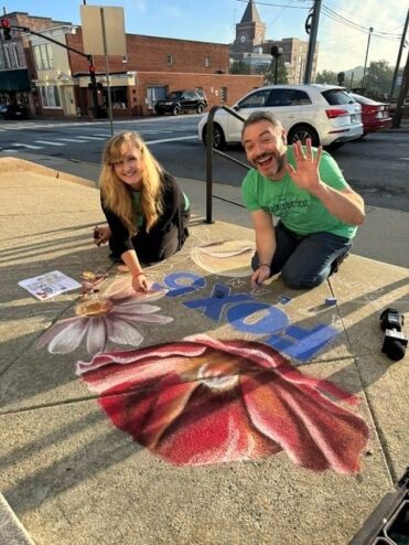 Jessi and Paul Milliken of FOX 5 Atlanta kneeling on the Marietta sidewalk beside the chalk drawing in progress, with the red poppy, a daisy and the FOX 5 logo, Paul waving
