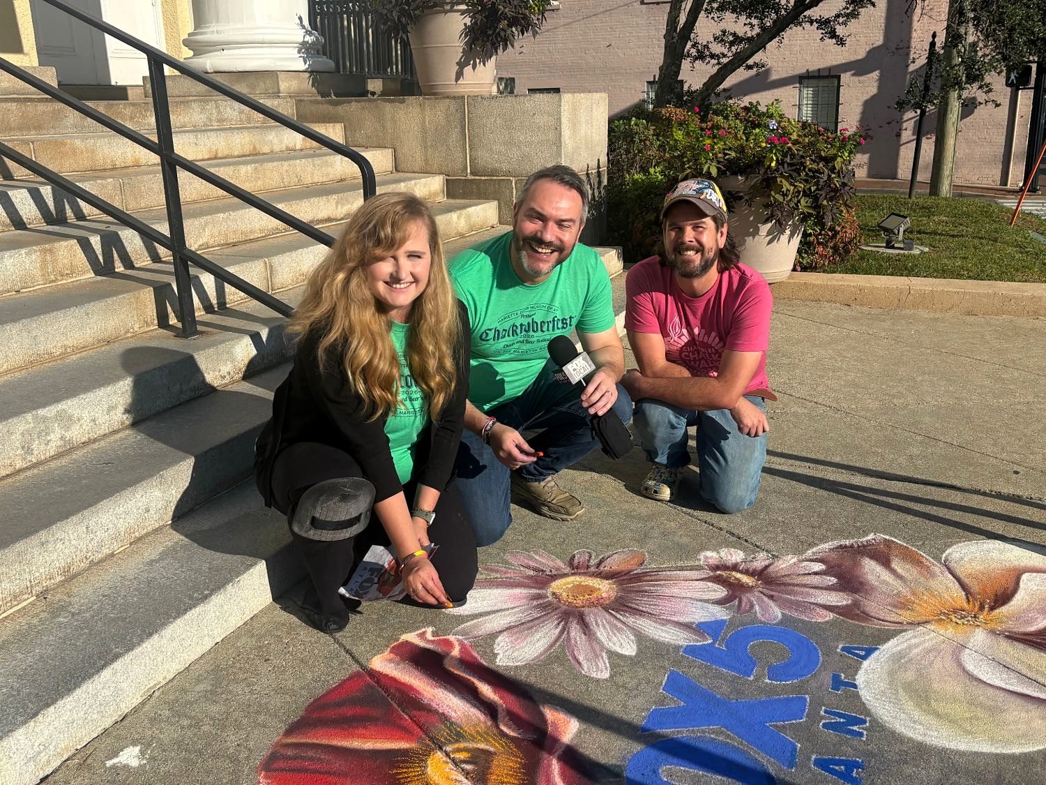 Jessi in a Chalktoberfest 2026 shirt kneeling with Paul Milliken from FOX 5 Atlanta, holding a FOX 5 microphone, and fellow chalk artist Zach Herndon beside the finished drawing