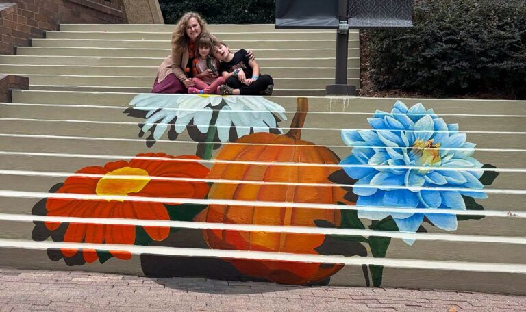 A mother and two children seated above a painted stair mural of a pumpkin between orange, white and blue flowers at Galleria on the Park
