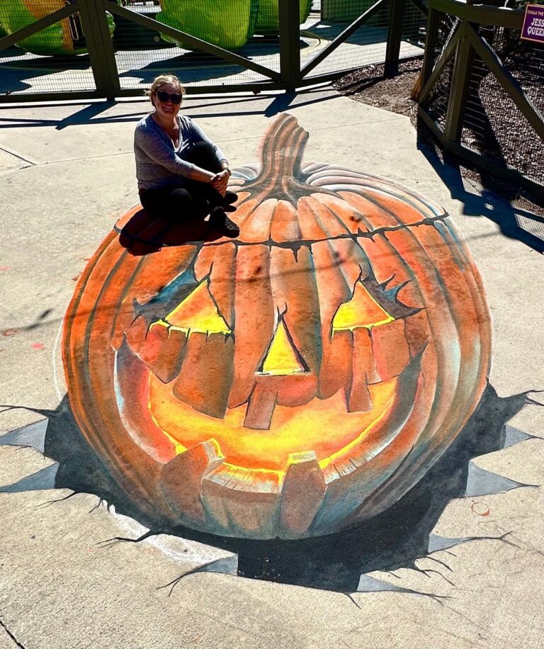 Jessi sitting on top of her giant 3D chalk jack-o'-lantern with a glowing carved grin, bursting up through the pavement
