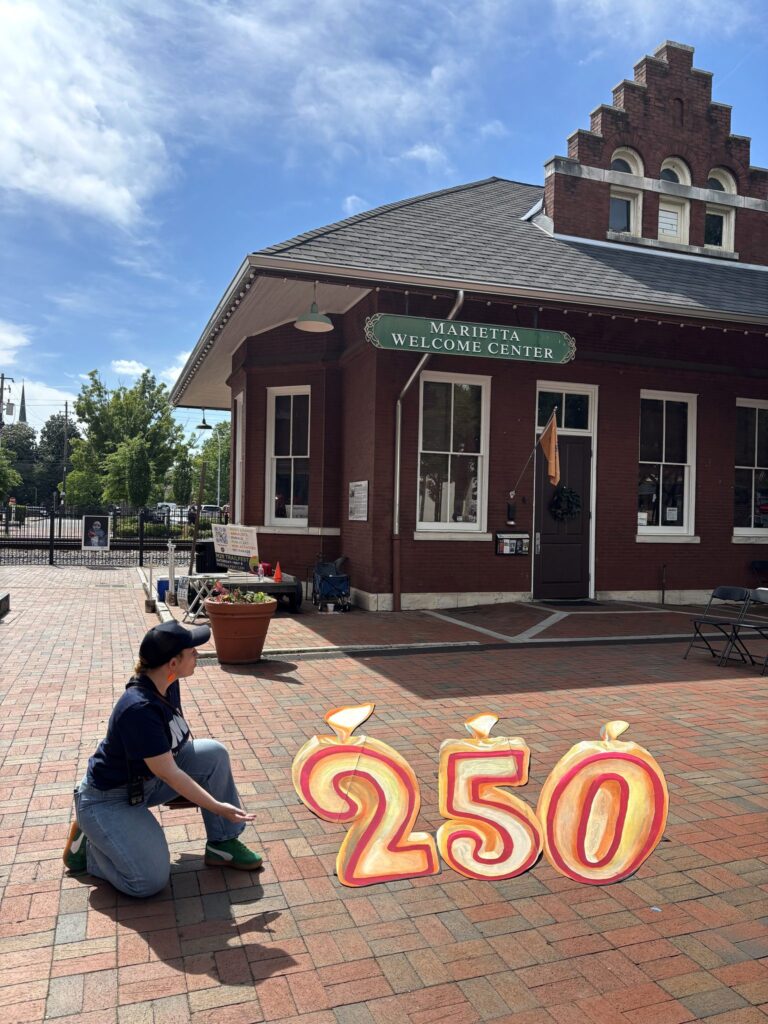 Artist kneeling beside chalk-painted 250 birthday candle numbers in front of the Marietta Welcome Center
