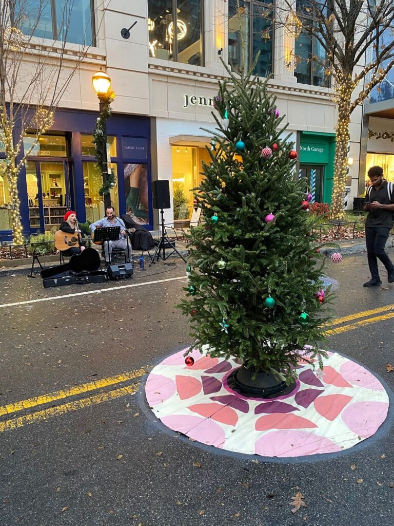 Christmas tree on a pink and plum petal-pattern painted tree skirt beside street musicians