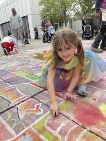 A smiling girl in rainbow fairy wings lying on the colorful chalk drawing, her hands covered in pastel chalk