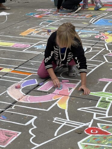 A visitor kneeling on the plaza filling in the chalk line drawing with pink, purple and yellow