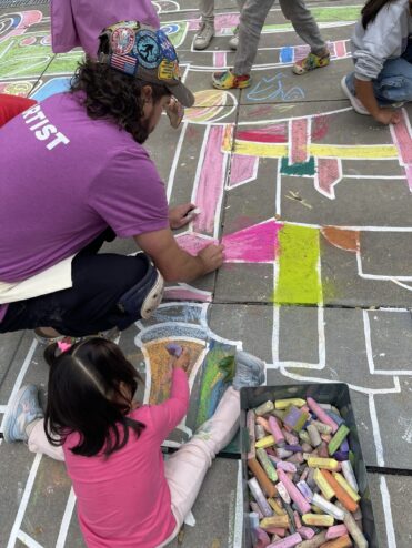 Zach in a purple Artist shirt coloring the drawing beside a little girl in pink, with a bin of chalk next to them