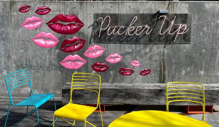 Mural of pink and red painted lips beside a Pucker Up neon sign, above colorful patio chairs